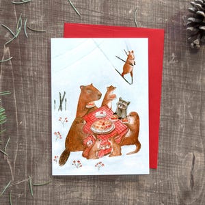Peut inclure: Carte de vœux fantaisiste avec une illustration à l'aquarelle d'animaux de la forêt réunis autour d'un pique-nique dans la neige. Une enveloppe rouge est derrière la carte. Un écureuil fait du ski en arrière-plan.