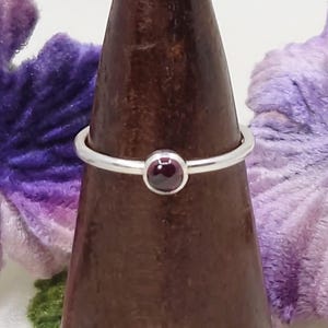 May include: A silver ring with a small, round, deep red gemstone. The ring is displayed on a brown, wooden ring holder against a backdrop of purple flowers. The gemstone is the focal point of the simple, elegant design.