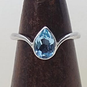 Könnte beinhalten: Ein silberner Ring mit einem tropfenförmigen, hellblauen Edelstein. Der Ring hat ein schlichtes, elegantes Band. Der Ring wird auf einem dunklen Holzständer präsentiert, um sein Design und seine Farbe hervorzuheben.