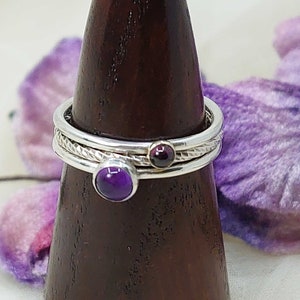 May include: Three silver rings with gemstones. The top ring features a purple amethyst, the middle ring has a red garnet, and the bottom ring has a textured band.