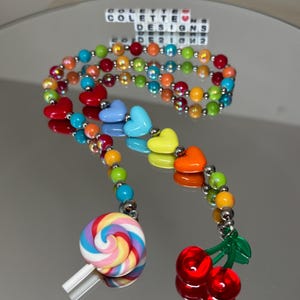 May include: Rainbow beaded necklace with heart charms, a lollipop, and a cherry charm. The necklace is made with colorful beads and silver spacers. The charms are made of plastic and are brightly colored.