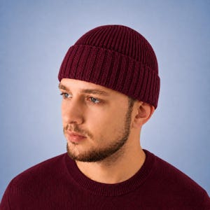 May include: A deep burgundy beanie crafted with a thick, vertical ribbed knit texture. The hat features a wide, folded cuff that sits securely around the head, providing a structured and uniform appearance in a solid, dark red color.