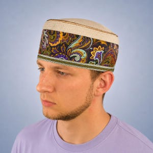 May include: A cylindrical kufi cap featuring a natural beige linen top and a wide band decorated with a vibrant paisley pattern in shades of purple, gold, green, and black, finished with a thin, detailed yellow and black woven trim at the base.