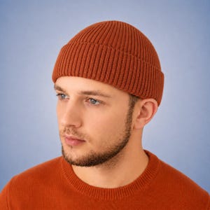 May include: A rust-colored beanie with a thick, vertical ribbed knit texture and a wide, folded cuff. The hat is constructed from a solid-colored yarn and features a rounded crown that fits closely to the head, providing a simple, uniform appearance.