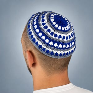 May include: A round, crocheted kippah featuring a concentric circular pattern in vibrant royal blue, crisp white, and soft grey yarn. The design consists of alternating bands of solid colours and intricate stitch work, creating a textured, layered appearance.