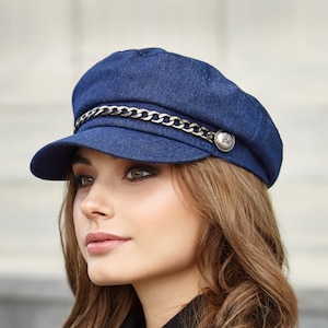 Op de afbeelding: Een blauwe denim newsboy-pet met een zilveren ketting en een zilveren knoop.