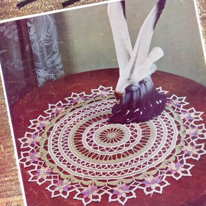 Puede incluir: Un encaje de ganchillo blanco con un diseño floral verde y rosa. El encaje está sobre una mesa roja con una figurita de pájaro blanco en el centro. El texto "A NEW LOOK in Crochet" está en la parte superior de la imagen. El texto "DESIGN BOOK No. 72" está en la parte inferior izquierda de la imagen. El texto "DIRECTIONS ON PAGE 15" está en la parte inferior derecha de la imagen. El texto "FAIR AND BAZAAR WINNERS" está en la parte inferior central de la imagen.