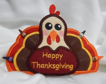 Puede incluir: Un porta lápices de fieltro con forma de pavo con una cola roja, naranja y amarilla. El pavo tiene un cuerpo blanco con una papada roja y un pico marrón. El porta lápices dice "Feliz Día de Acción de Gracias" en bordado dorado.