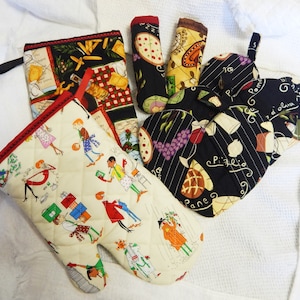 May include: Four quilted oven mitts with different Italian-themed designs. One mitt is cream with red trim and features a pattern of people carrying various items. Another mitt is black with red trim and features a pattern of grapes, bread, and the words "Pane" and "Pizzeria". The other two mitts are black with red trim and feature a pattern of pizza slices, tomatoes, and the words "Pomodoro" and "d'oliva".