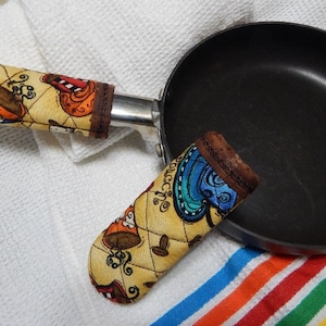 May include: Two colorful fabric pot holders with a coffee cup design. The pot holders are on a white towel with a rainbow striped towel underneath.