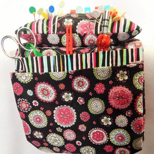 May include: A black and white fabric sewing organizer with a pincushion on top. The organizer has multiple pockets and a striped fabric lining. The pincushion is filled with colorful pins and needles.