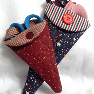 Puede incluir: Dos porta tijeras de tela con patrones rojo, blanco y azul. Los porta tijeras tienen forma de cono y tienen un bolsillo para tijeras. Un porta tijeras tiene un patrón de cuadros rojos y blancos y el otro tiene un patrón de rayas rojas, blancas y azules. Ambos porta tijeras tienen un patrón de estrellas azules en la parte posterior.