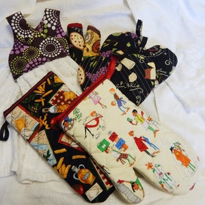 May include: Set of four kitchen oven mitts. One is black with white and red accents and has the words "Pizzello" and "Pomodoro" printed on it. Another is black with white and red accents and has a pattern of pasta and olive oil bottles. Another is cream with black and red accents and has a pattern of people and food. The last one is purple with white and green accents and has a circular pattern.