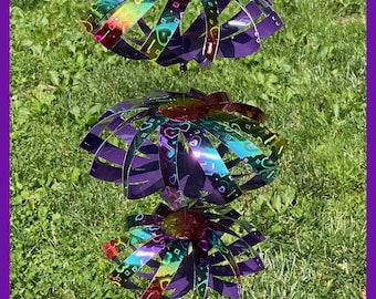 May include: A hanging wind spinner featuring three stacked, circular tiers made of iridescent, rainbow-colored material with a repeating heart pattern. The structure is connected by a thin wire and topped with a single purple bead.
