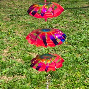 May include: A pink and iridescent wind spinner with three tiers. Each tier is a circular shape with a design of overlapping strips. The wind spinner is hanging from a string in a grassy area.