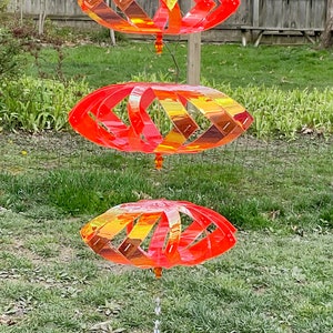 May include: A red and orange wind spinner with three tiers of overlapping, diamond-shaped panels. The spinner is suspended from a crystal-shaped pendant.