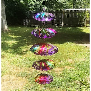 Könnte beinhalten: Ein hängendes Windspiel mit vier irisierenden, geometrischen Formen in Lila, Rosa und Grün. Die Formen sind an einer transparenten Schnur aufgehängt und bilden einen dekorativen Akzent im Freien. Das Windspiel befindet sich vor dem Hintergrund von grünem Gras und Bäumen.
