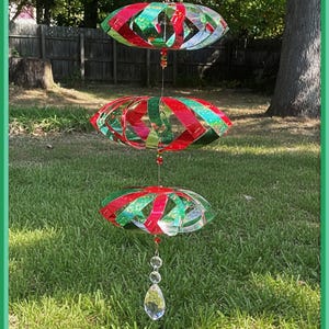 May include: A three-tiered, hanging outdoor decoration with red and green glitter accents. The decorative pieces are oval-shaped and suspended by a string, with a clear crystal pendant at the bottom. The background features green grass and trees.