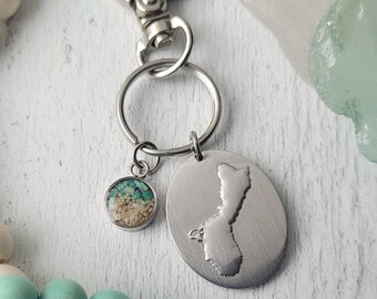 May include: A silver keychain with a round charm filled with sand and turquoise stones, and a larger oval pendant with a state outline. The keychain has a clasp and a ring. Wooden beads in cream and turquoise are in the background.