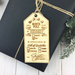 May include: A wooden Christmas gift tag with a North Pole delivery label. The tag is decorated with reindeer and has a checklist for naughty or nice, delivery date, and delivery method. The tag is addressed to Kennedy and signed by Santa Claus.