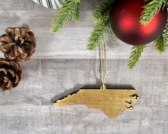 May include: A wooden ornament shaped like the state of North Carolina, hanging from a string with a red Christmas bauble and pine branches in the background.