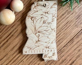 May include: A wooden Mississippi state ornament with a floral design and the state name engraved at the bottom. The ornament is light brown with a natural wood grain and a jute string for hanging. The ornament is surrounded by Christmas decorations.