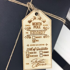 May include: A wooden Christmas gift tag with a North Pole Express theme. The tag is decorated with a reindeer and sleigh, and has a checklist for Santa Claus to fill out. The tag is addressed to Kennedy and includes a delivery date of December 25th. The tag is from Santa Claus.