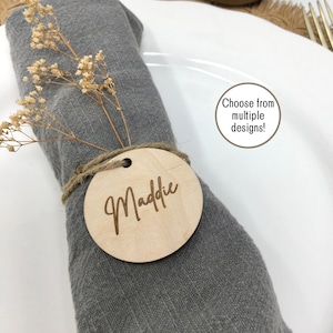 May include: A round wooden place card with the name "Maddie" engraved on it. The place card is tied to a gray napkin with twine. The place card is on a white plate with a text overlay that says "Choose from multiple designs!"