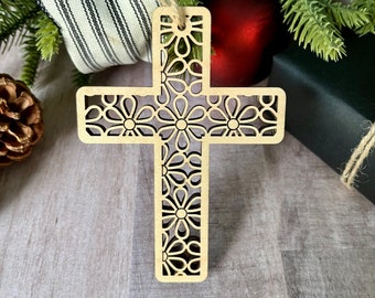 May include: A wooden cross ornament with a floral design. The cross is light brown and has a delicate, intricate pattern of flowers cut out of the wood.