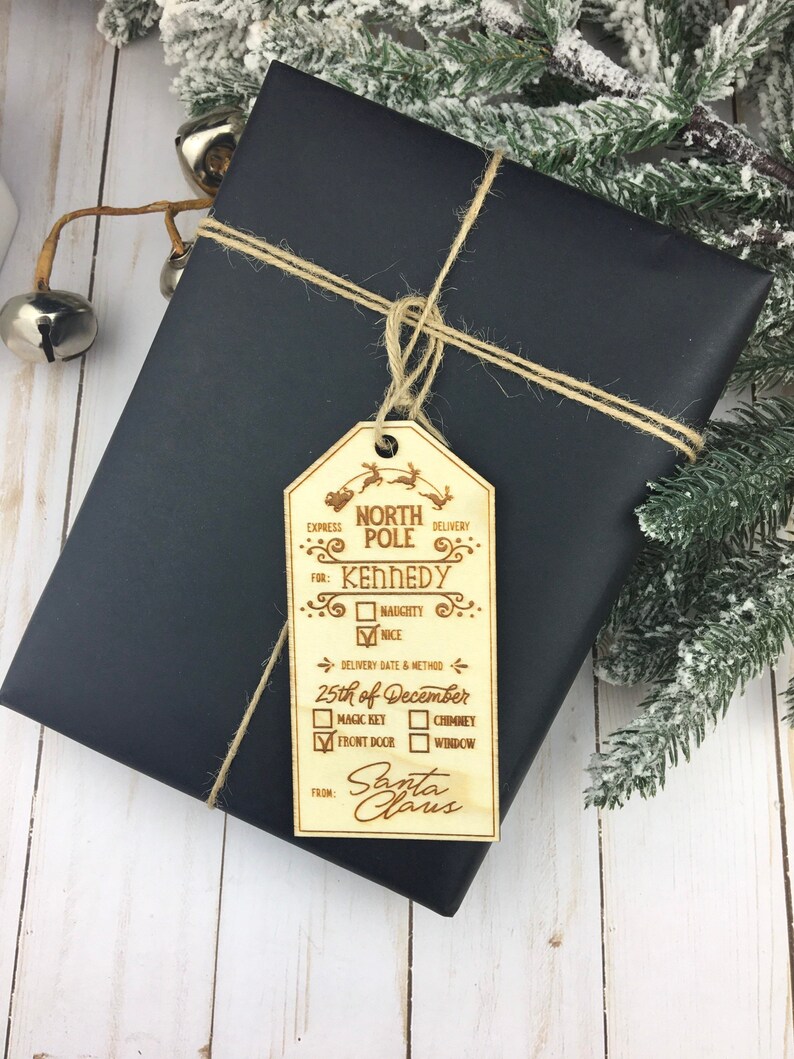 May include: A wooden Christmas gift tag with a North Pole delivery label. The tag is decorated with reindeer and has a checklist for naughty or nice, delivery date, and delivery method. The tag is addressed to Kennedy and signed by Santa Claus.