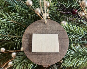 May include: A wooden Christmas ornament shaped like a circle with a smaller square cutout in the center. The ornament is painted a dark brown color with a light brown square cutout. The ornament is hanging from a string.