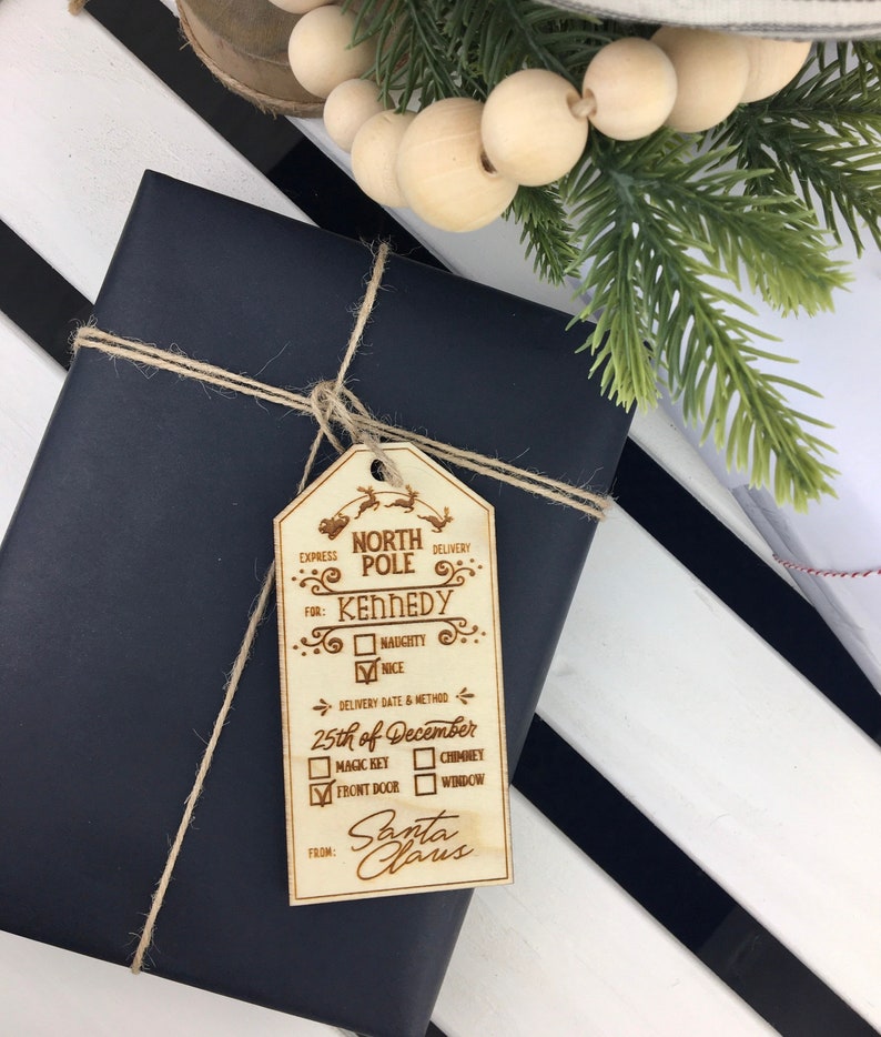 May include: A wooden Christmas gift tag with a North Pole theme. The tag is decorated with a reindeer and sleigh design. The tag is personalized with the name "Kennedy" and includes a delivery date of December 25th. The tag also includes options for delivery method, including magic key, chimney, front door, and window. The tag is signed "Santa Claus".