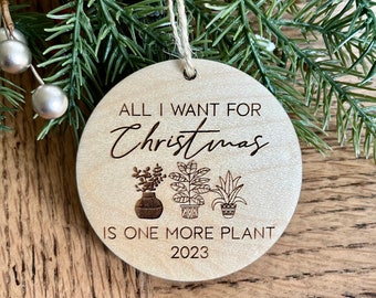 May include: A round wooden Christmas ornament with the text "All I want for Christmas is one more plant 2023". The ornament features three stylized potted plants.