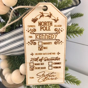 May include: A wooden Christmas ornament shaped like a tag with the text "North Pole Delivery" and "Kennedy" with checkboxes for "Naughty" or "Nice". The ornament also includes a delivery date and method section with checkboxes for "Magic Key", "Chimney", "Front Door", and "Window". The ornament is signed "Santa Claus".