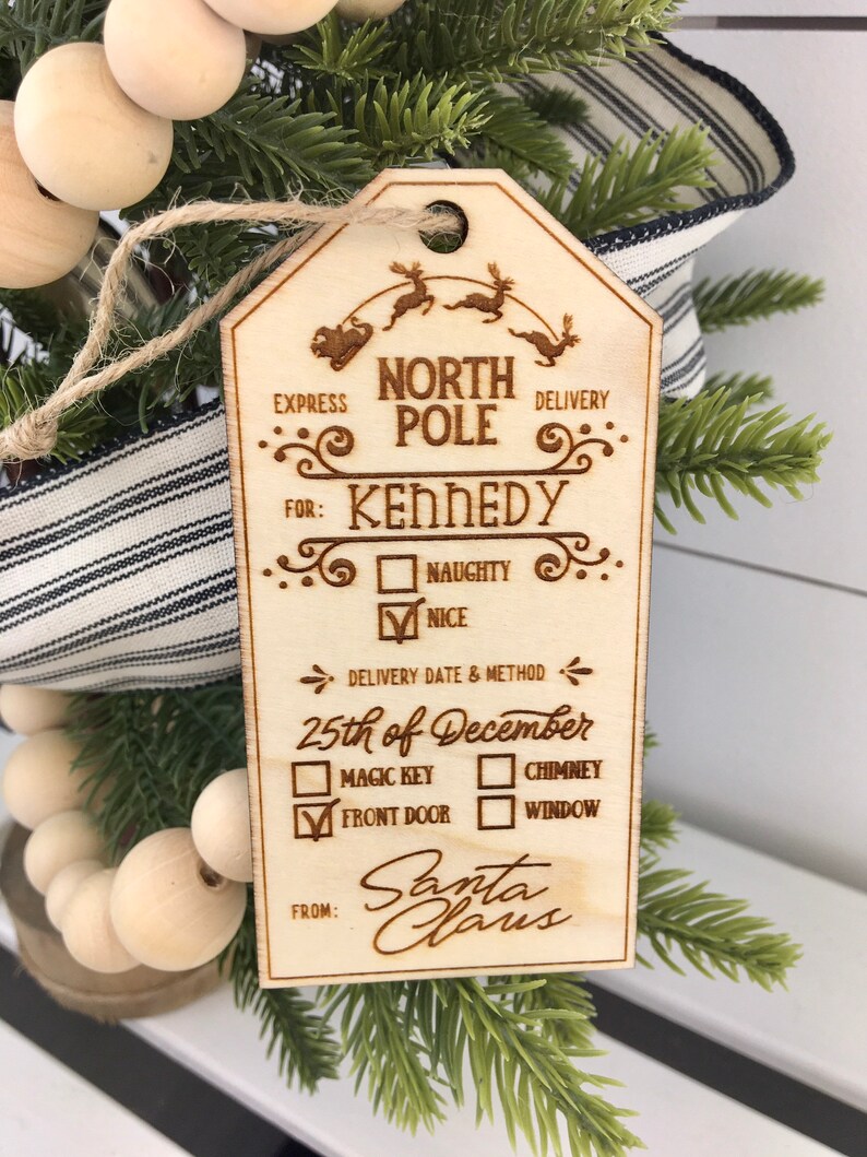 May include: A wooden Christmas ornament shaped like a tag with the text "North Pole Delivery" and "Kennedy" with checkboxes for "Naughty" or "Nice". The ornament also includes a delivery date and method section with checkboxes for "Magic Key", "Chimney", "Front Door", and "Window". The ornament is signed "Santa Claus".