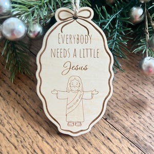 May include: A light-colored wooden ornament with a scalloped edge and a jute twine hanger. The text reads "EVERYBODY NEEDS A LITTLE Jesus" above a line-art illustration of a figure with arms outstretched, wearing a sash that says "Jesus loves you".