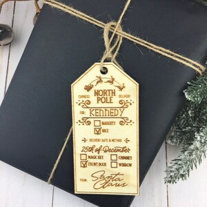 May include: A wooden Christmas gift tag with a North Pole theme. It has a space for the recipient's name, a naughty or nice checkbox, a delivery date and method, and a space for Santa Claus to sign. The tag is tied with twine to a black gift box.
