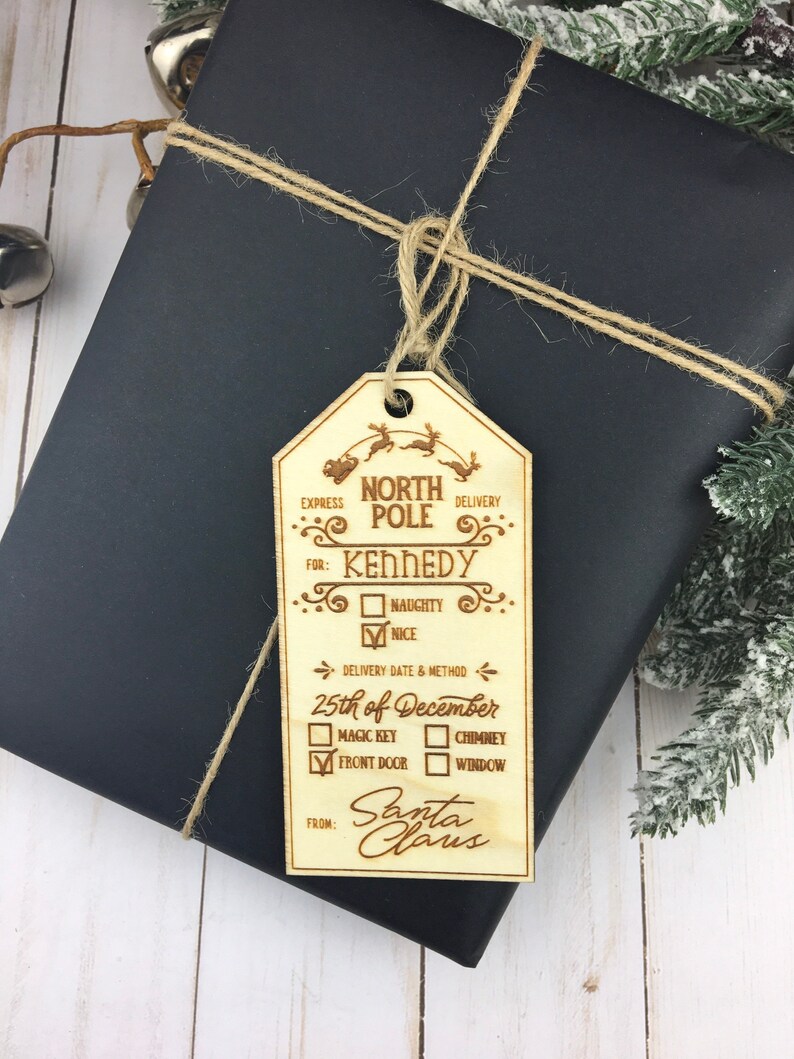 May include: A wooden Christmas gift tag with a North Pole theme. It has a space for the recipient's name, a naughty or nice checkbox, a delivery date and method, and a space for Santa Claus to sign. The tag is tied with twine to a black gift box.