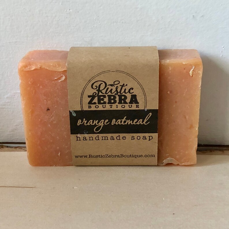 May include: A rectangular bar of orange oatmeal handmade soap with a brown paper label. The label features the text "Rustic Zebra Boutique" and "orange oatmeal handmade soap". The soap is on a white surface.