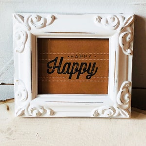 Op de afbeelding: Witte, sierlijke fotolijst met een bruine achtergrond en de tekst "HAPPY. Happy" in zwarte letters.