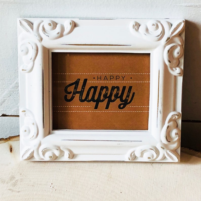 Op de afbeelding: Witte, sierlijke fotolijst met een bruine achtergrond en de tekst "HAPPY. Happy" in zwarte letters.