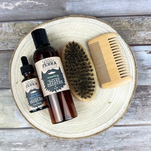 May include: A set of beard care products on a wooden surface. The set includes a bottle of beard shampoo labeled "woodsy", a bottle of beard oil labeled "texas cedar lime", a wooden comb, and a boar bristle brush.