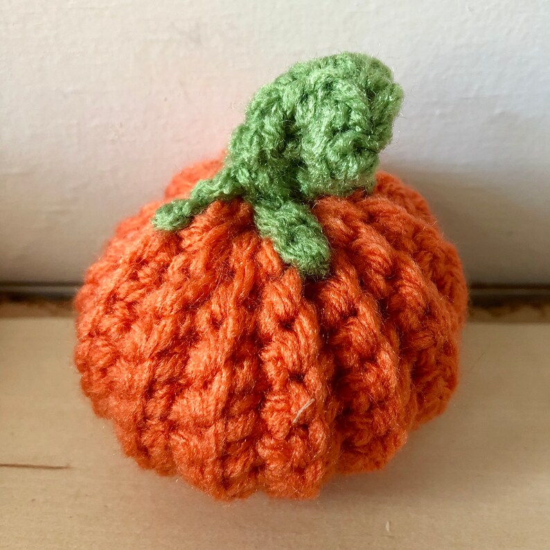 May include: A handmade, orange crochet pumpkin with a green stem. The pumpkin is round with ribbed detailing, and the stem is a cluster of green leaves. This decorative item is perfect for fall and autumn-themed decor.
