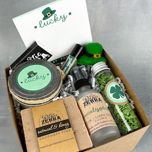 May include: A gift box filled with green and brown themed items for St. Patrick's Day. The box includes a candle, soap, a bottle of essential oil, a small bottle of green sprinkles, and a green felt leprechaun hat. The box is decorated with a green shamrock and the text "*lucky*"