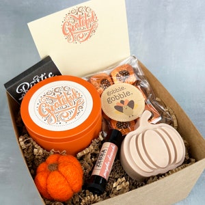 May include: A brown gift box filled with a variety of Thanksgiving-themed items, including a candle, chocolates, a lip balm, and a felt pumpkin. The box is decorated with brown paper and shredded paper. The candle is orange and has the word "grateful" written on it. The chocolates are wrapped in gold foil and have a "gobble, gobble" sticker on them. The lip balm is in a small tube and has a pumpkin spice scent. The felt pumpkin is orange and has a green stem.