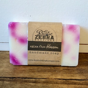 May include: A bar of handmade soap with a white base and pink swirls. The soap is labeled "asian tree blossom" and is from the Rustic Zebra Boutique.