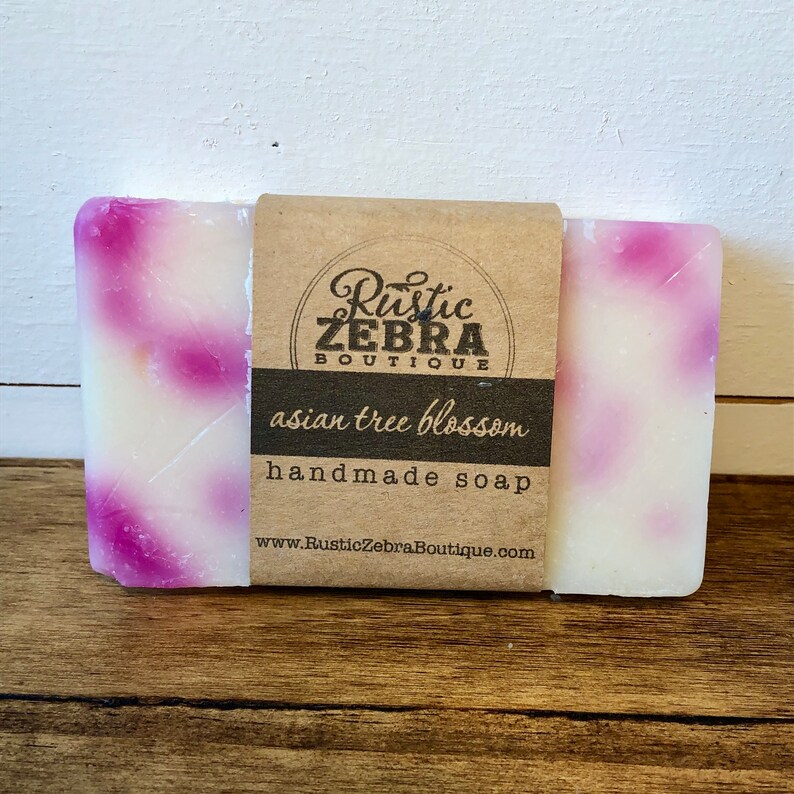 May include: A bar of handmade soap with a white base and pink swirls. The soap is labeled "asian tree blossom" and is from the Rustic Zebra Boutique.