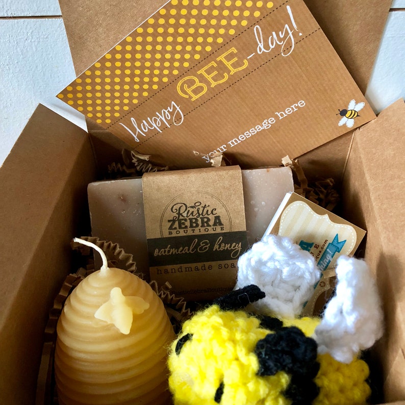 May include: A gift box filled with bee-themed items, including a honey-scented candle shaped like a honeycomb, a crocheted bee toy, a bar of oatmeal and honey soap, and a card that says "Happy Bee-day!"
