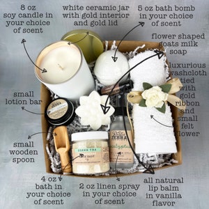 May include: A gift box filled with bath and body products, including an 8 oz soy candle, a 5 oz bath bomb, a small lotion bar, a small wooden spoon, a white ceramic jar with a gold lid, a flower-shaped goats milk soap, a luxurious washcloth tied with a gold ribbon, a 2 oz linen spray, and a 4 oz bath bomb. The box is filled with shredded paper and the products are arranged in a visually appealing way.