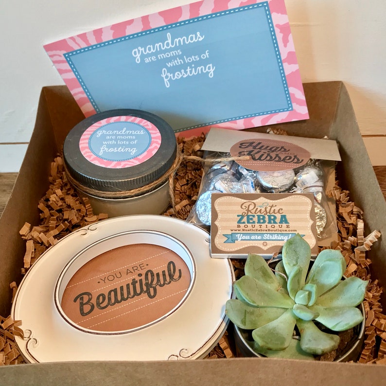 May include: A gift box filled with a variety of items, including a candle, a succulent plant, a small plate with the words "You are Beautiful" on it, and a bag of chocolate kisses. The box is lined with brown paper shreds. The top of the box has a card with the text "Grandmas are moms with lots of frosting".